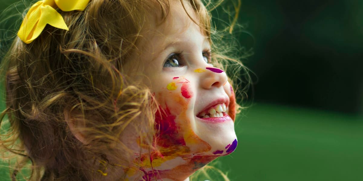 Smiling toddler covered in colorful paint splatters, wearing a yellow hair bow, outdoors