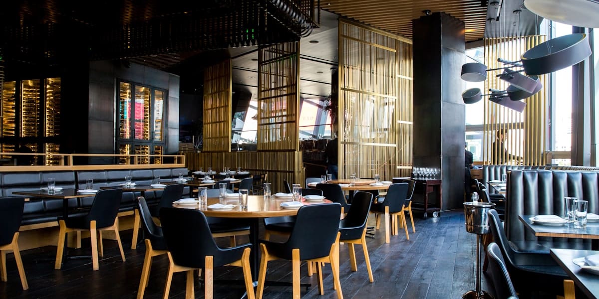 A modern upscale restaurant dining room with round wooden tables, black chairs, a backlit wine display wall and pendant lighting.